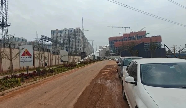Perspective view of the access road flanked by parked cars, with completed high-rise apartments and ongoing construction visible in the distance