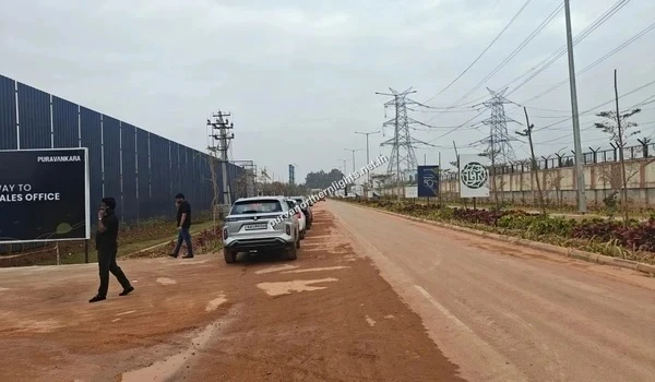 Wide view of the entrance road leading to the Puravankara sales office, featuring blue site hoarding, parked vehicles, and directional signage