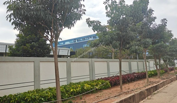 View of the RTX building rising above a long compound wall lined with trees and colorful garden plants along the road near Purva Northern Lights