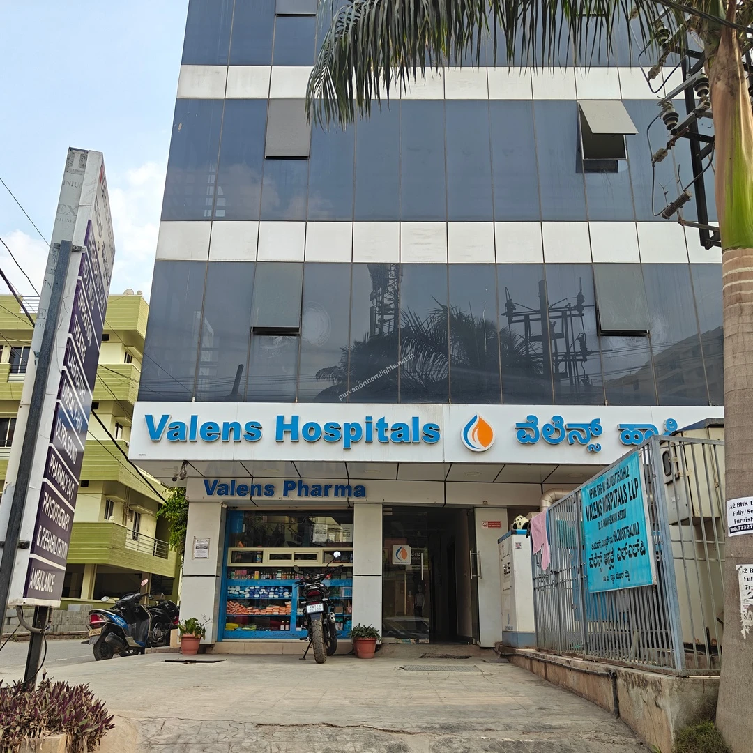 The modern glass exterior of Valens Hospitals seen on a bright Mar 13 afternoon highlighting premium healthcare options near Purva Northern Lights.