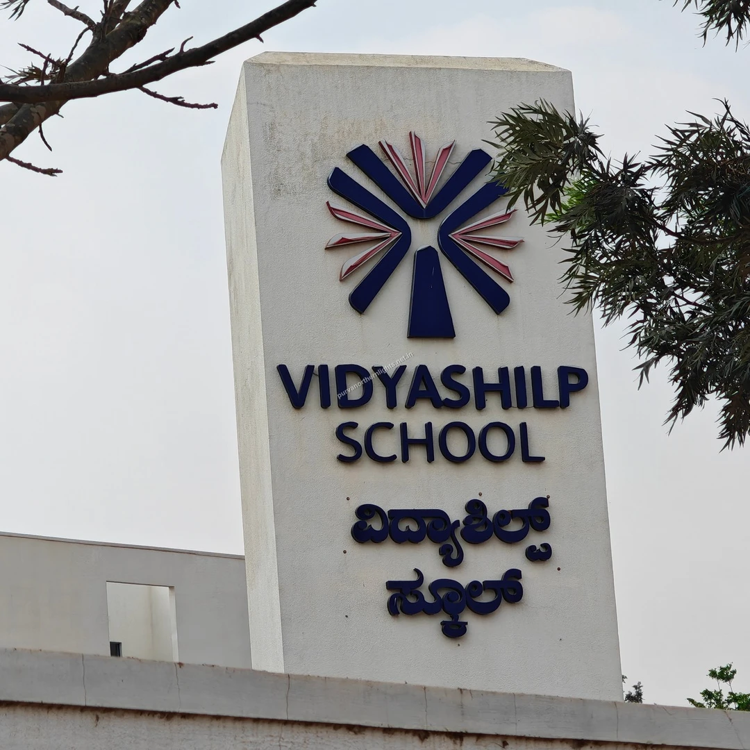 Vidyashilp Signage Trees The white Vidyashilp School landmark pillar standing tall next to leafy green branches on a clear Mar 13 morning.
